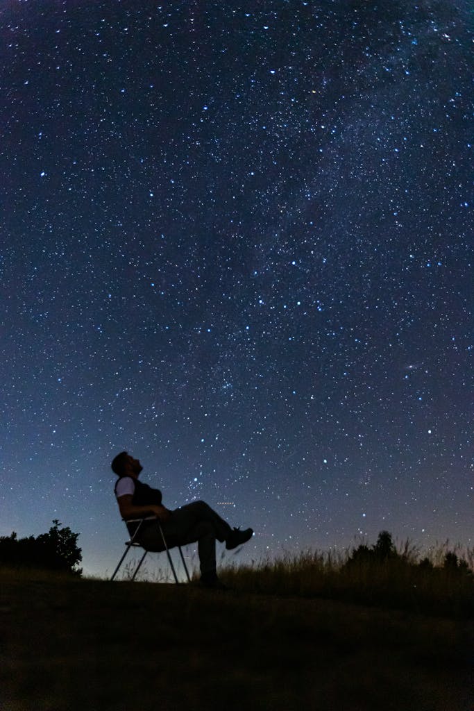 A man sits quietly on a hill, silhouetted against a vast starry night sky in Bosnia.